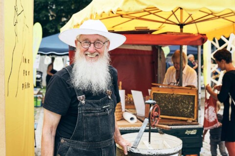 Ein fröhlicher älterer Mann mit weißem Hut und Bart bei einem Marktstand in Stuttgart.