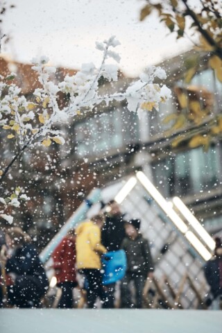 Blick durch ein Fenster mit Schneeflocken auf eine Gruppe von Menschen im Freien vor einem modernen.