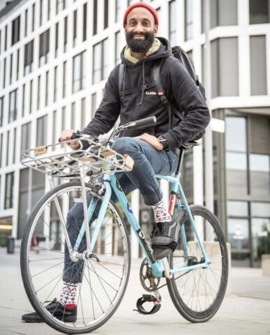 Ein fröhlicher Mann mit Rucksack auf Fahrrad vor moderner Stadtarchitektur.