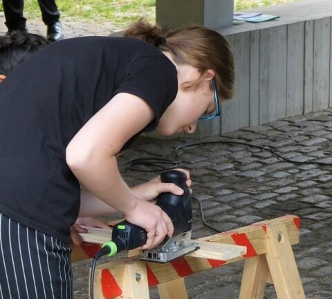 Jugendliche beim Holzarbeiten mit Stichsäge auf Baustelle im Freien.