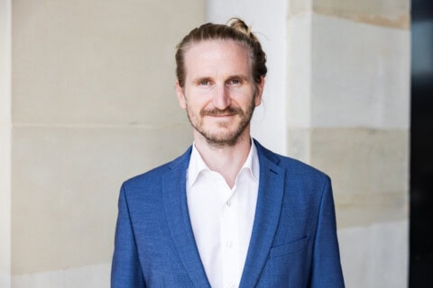 Ein Mann im blauen Anzug vor einer Steinwand, professionell und freundlich.