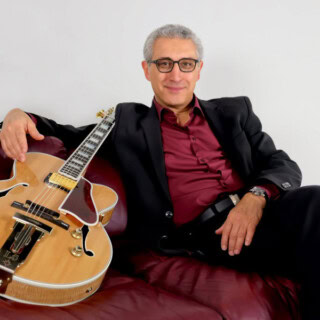 Ein Mann mit Brille hält eine Gitarre und sitzt auf einem roten Sofa.