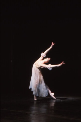 Elegante Balletttänzerin in weißem Kostüm auf dunklem Bühnenhintergrund.