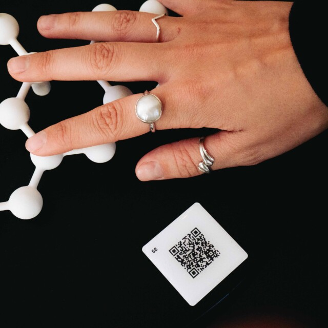 Eine Hand mit Ringen hält eine Molekülmodell-Struktur, QR-Code liegt daneben.