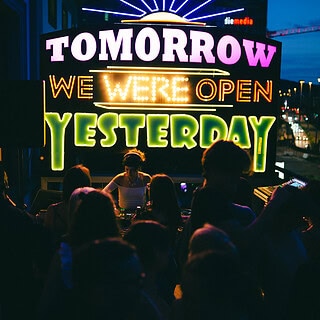 Neon-Schriftzug mit 'Tomorrow', 'We Were Open' und 'Yesterday' vor einer Menschenmenge bei Nacht.
